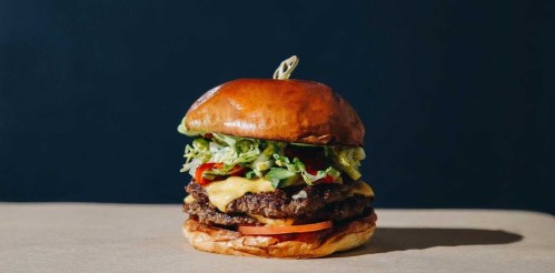 A tall burger with a shiny bun, layers of lettuce, tomato, cheese, and two beef patties, set against a dark background.