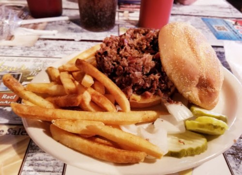 A plate with a pulled meat sandwich, fries, pickles, and onions on a table.