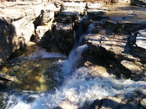 A small waterfall cascades over rocky terrain, surrounded by sunlight and clear water.