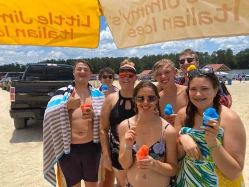 A group of six people at the beach, smiling and holding colorful ice treats under a sunny sky.