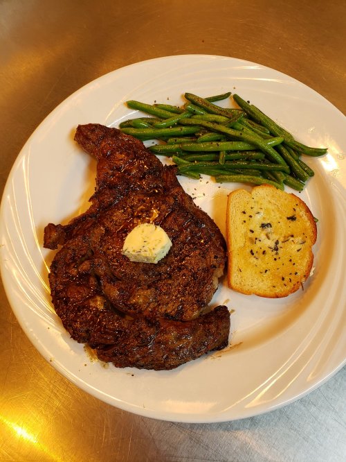 A plate with a grilled steak topped with butter, served with green beans and a slice of garlic bread.