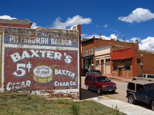 A vintage mural advertising Baxter's 5¢ Cigar on a brick wall, with historic buildings and cars in a small town setting.