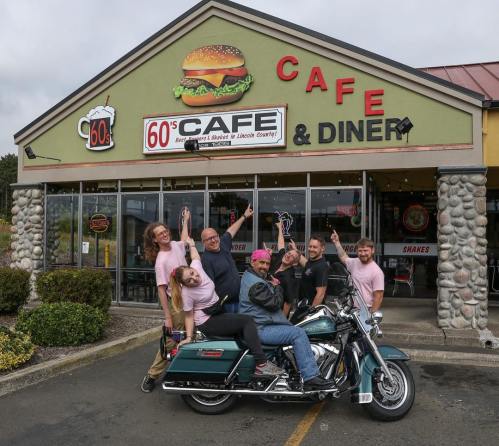 A group of six people poses playfully around a motorcycle in front of a retro diner with a large burger sign.