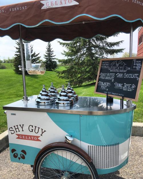A gelato cart with various flavors displayed on a chalkboard, set against a green landscape in Vermont.