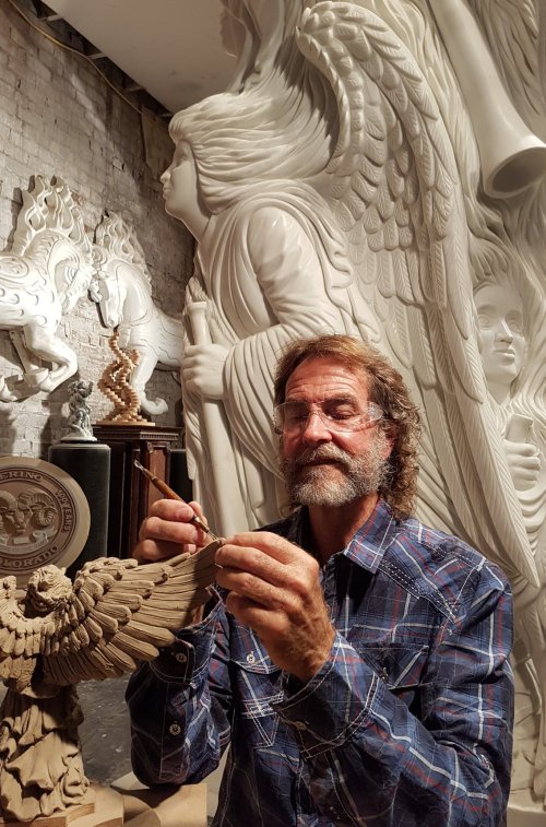 An artist carves a detailed sculpture of an angel, surrounded by other intricate sculptures in a studio.