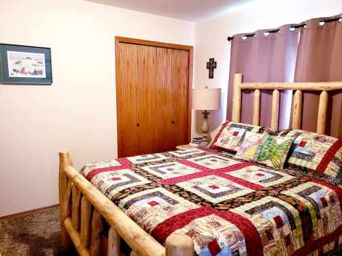 Cozy bedroom featuring a log bed, colorful quilt, lamp, and wooden closet, with curtains and a framed picture on the wall.