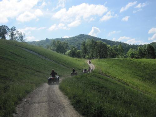 ATVs riding on a dirt path through green hills under a blue sky with fluffy clouds.