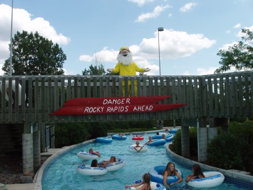 A water park scene with a yellow-clad figure warning of "Danger: Rocky Rapids Ahead" above a lazy river with floating guests.