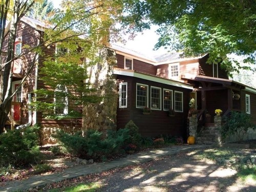 A charming brown wooden house surrounded by trees, featuring a stone chimney and a welcoming front porch.