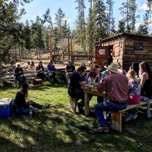 A group of people enjoying a meal outdoors at a rustic setting with trees and a wooden cabin in the background.