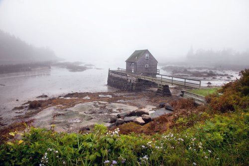 A foggy landscape featuring a small house on stilts by a calm, shallow waterway surrounded by rocky terrain and greenery.