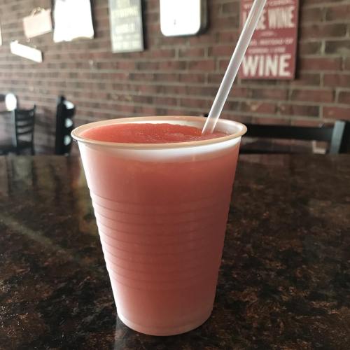 A pink frozen drink in a clear plastic cup with a straw, sitting on a dark countertop in a rustic setting.