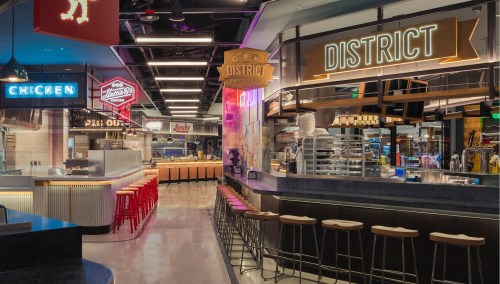 A modern food hall with vibrant signage, featuring a bar area and various dining options, illuminated by neon lights.