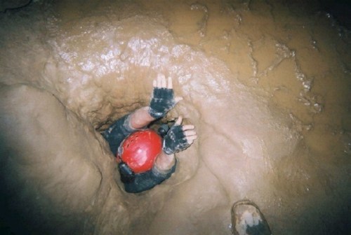 A person in a red helmet reaches up from a narrow cave passage, surrounded by wet, muddy walls.