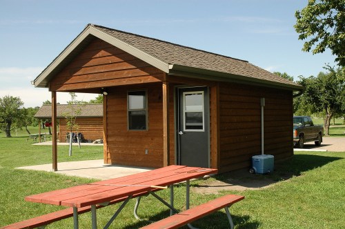 A small wooden cabin with a porch, surrounded by grass and a picnic table in front. A truck is parked nearby.
