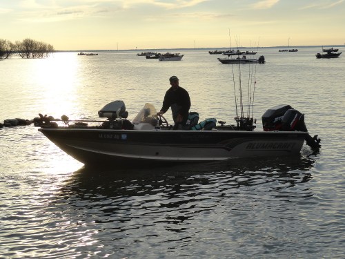 A man stands on a fishing boat in calm waters, with several boats visible in the background under a sunset sky.