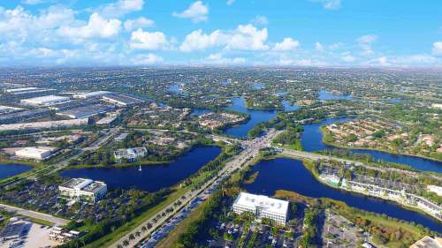 Aerial view of a lush landscape with lakes, roads, and suburban areas under a bright blue sky with scattered clouds.