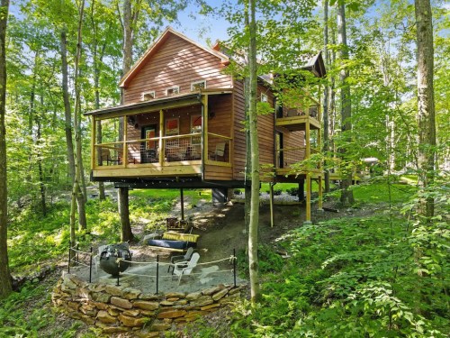 A modern wooden cabin elevated on stilts, surrounded by lush green trees and a stone patio with outdoor seating.
