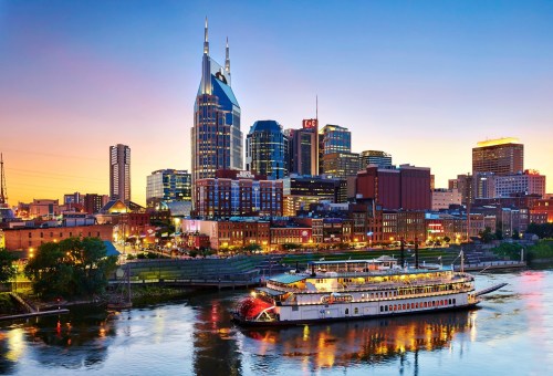 A scenic view of Nashville's skyline at sunset, featuring a riverboat on the water and illuminated buildings.
