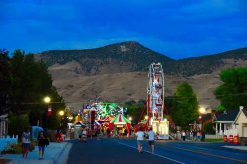 A lively carnival scene at dusk, with a Ferris wheel and colorful lights, set against a mountainous backdrop.