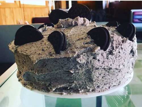 A round cake with gray frosting and topped with Oreo cookies, displayed on a glass surface.