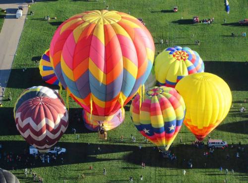 Aerial view of colorful hot air balloons on a grassy field, with people gathered around them.