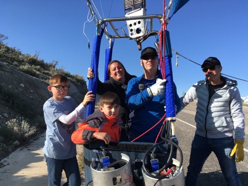 A group of five people poses beside a hot air balloon basket on a sunny day, smiling and enjoying the moment.