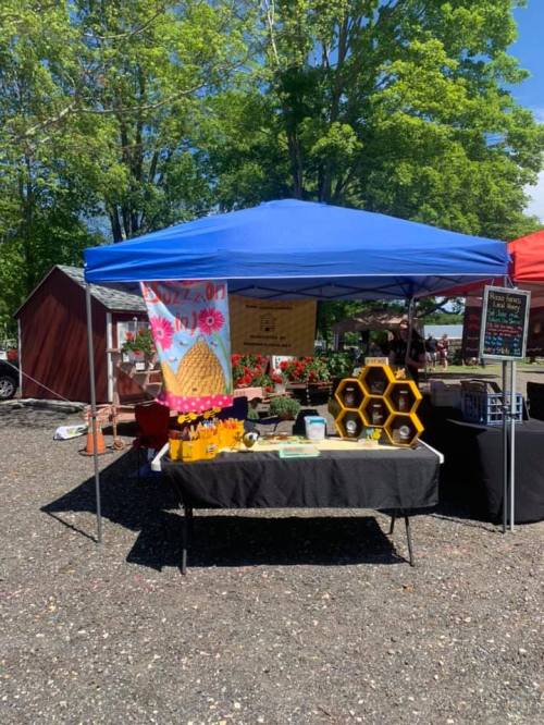 A blue tent at a market with honey products displayed on a table, surrounded by greenery and colorful flowers.