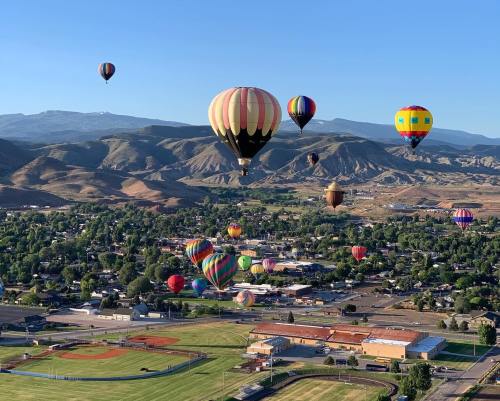 A vibrant scene of colorful hot air balloons floating over a town and green fields, with mountains in the background.