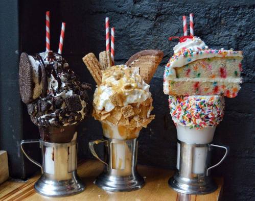 Three extravagant milkshakes topped with whipped cream, cookies, sprinkles, and cake slices, served in metal cups.