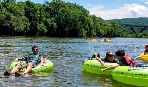 A man relaxes on a tube in a river, while two kids chat on another tube, with others floating in the background.