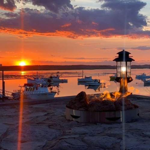 A serene sunset over a calm harbor, with boats and a lantern beside a small fire pit.