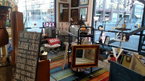 A cozy shop interior filled with vintage decor, signs, and colorful rugs, with a large window showing the street outside.