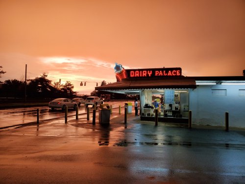 A retro ice cream shop, "Dairy Palace," illuminated against a dramatic orange sunset, with wet pavement reflecting the light.