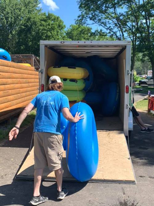 A person in a blue shirt loads colorful inflatable tubes into a trailer on a sunny day.