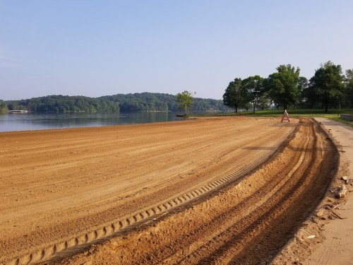 A sandy shoreline by a calm lake, with a person walking along a freshly graded path and trees in the background.