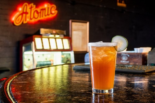 A refreshing drink with ice and a cucumber slice sits on a bar counter, with a neon "Atomic" sign in the background.