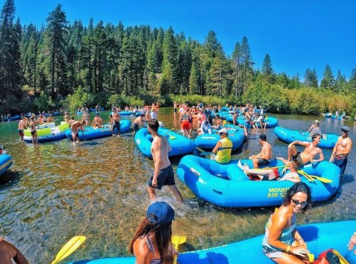 A lively scene of people enjoying a sunny day on blue rafts in a river surrounded by trees.