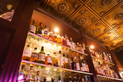 A well-stocked bar shelf displaying various bottles of liquor under warm lighting and ornate ceiling details.