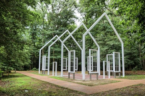 A white, abstract sculpture resembling houses with open frames and windows, set in a lush green forest.
