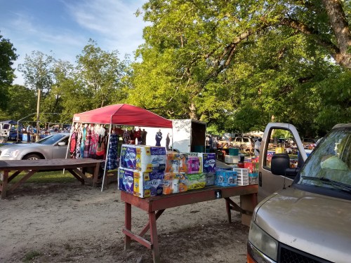 A vibrant outdoor market scene with a red tent, various goods for sale, and parked vehicles under green trees.