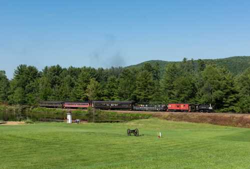 A vintage train travels along a scenic route beside a pond, surrounded by lush green trees and hills.