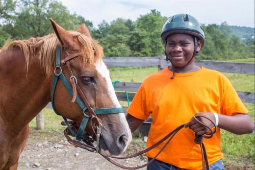 A smiling child in an orange shirt holds the reins of a brown horse, set against a green landscape.