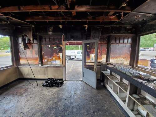 Interior of a burned building with charred walls, a damaged ceiling, and open doors revealing a smoky exterior.