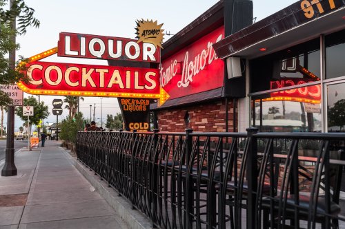 Neon signs for "Liquor" and "Cocktails" illuminate a sidewalk outside a bar, with a fence lining the patio area.