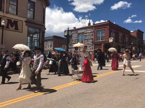 A lively street scene with people in vintage clothing, some carrying parasols, during a parade on a sunny day.