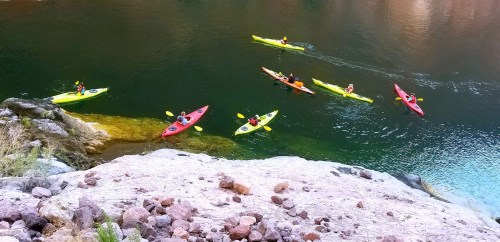 Aerial view of kayakers in colorful kayaks paddling on a calm river surrounded by rocky cliffs.