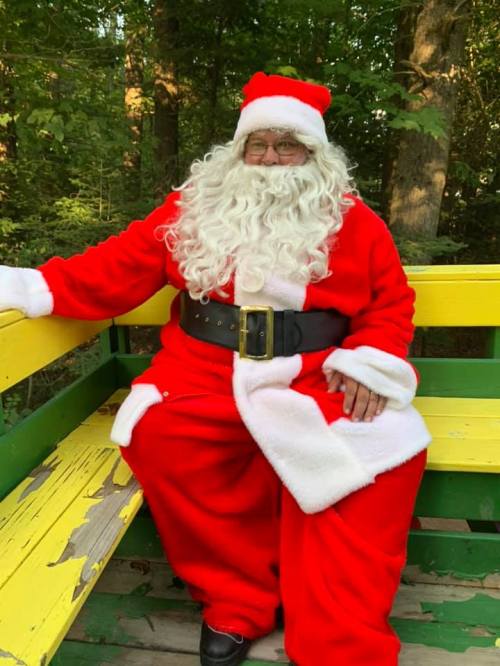 A person dressed as Santa Claus sits on a colorful bench in a wooded area, wearing a red suit and white beard.