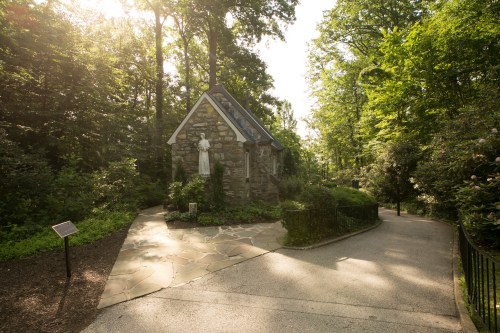 A stone chapel surrounded by lush greenery, with a statue and a pathway leading through the serene landscape.