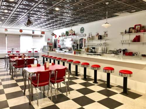 A retro diner interior with red stools, checkered floor, and a counter, featuring vintage decor and a welcoming atmosphere.